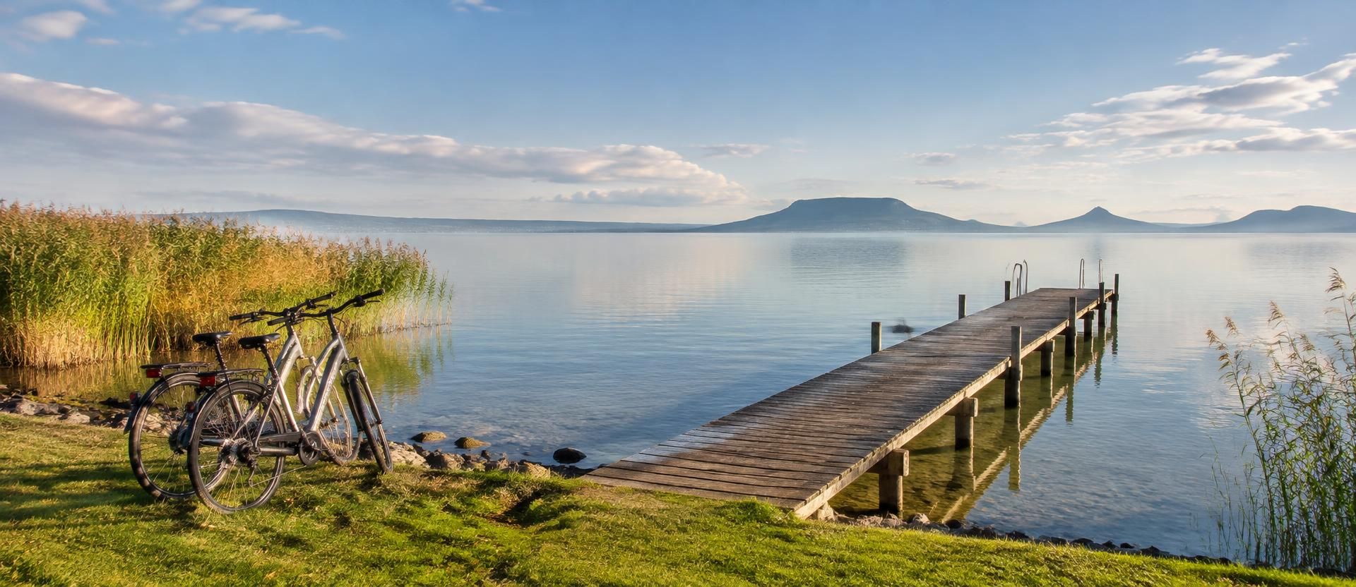 Fából készült stég a Balatonon, kerékpárok a füves parton, háttérben a tanúhegyekkel